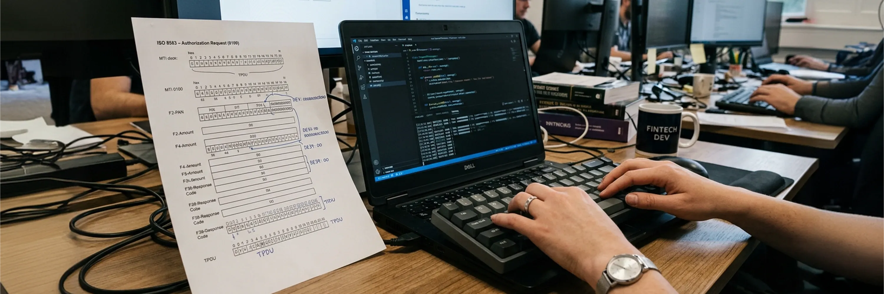 Fintech developer coding payment processing software on a laptop beside ISO 8583 transaction authorization documentation in a modern office workspace
